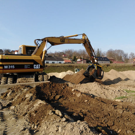 Caterpillar M315 excavator digging in a construction site with houses in the background.