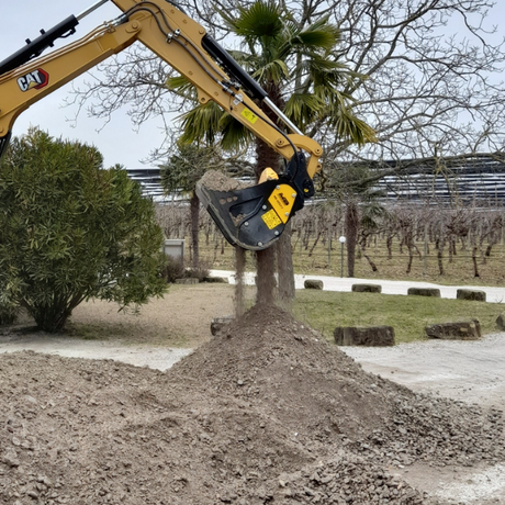 MB HDS212 Excavator arm with a tree stump removal tool in an outdoor setting with trees and a fence.