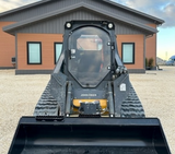 A John Deere skid steer with a clear polycarbonate door installed on it, front view with a building in the background.